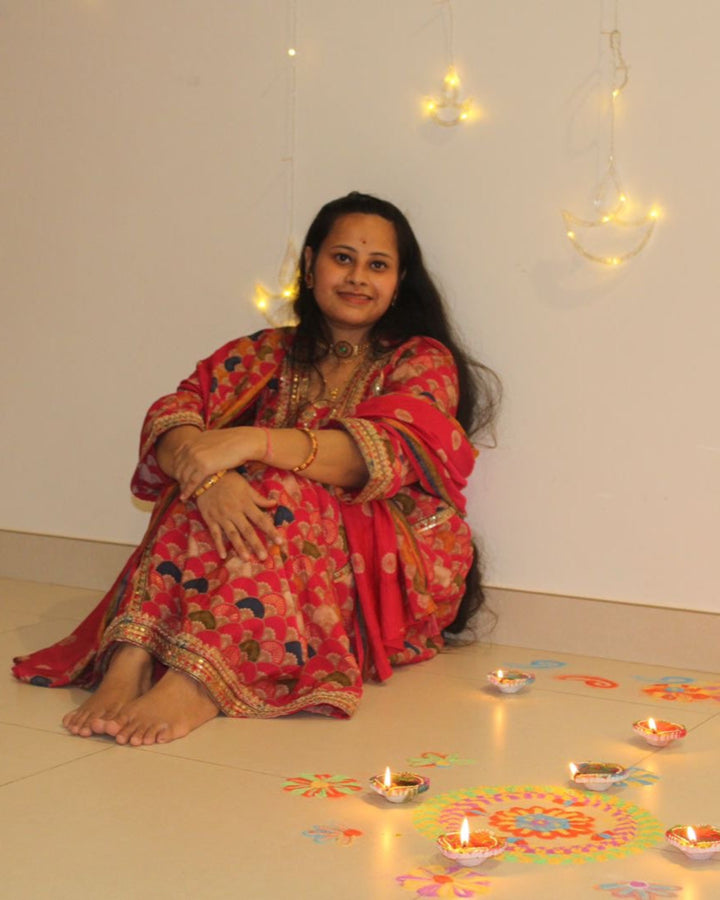 Woman in a red suit sitting on the floor with decorative oil lamps and rangoli patterns.