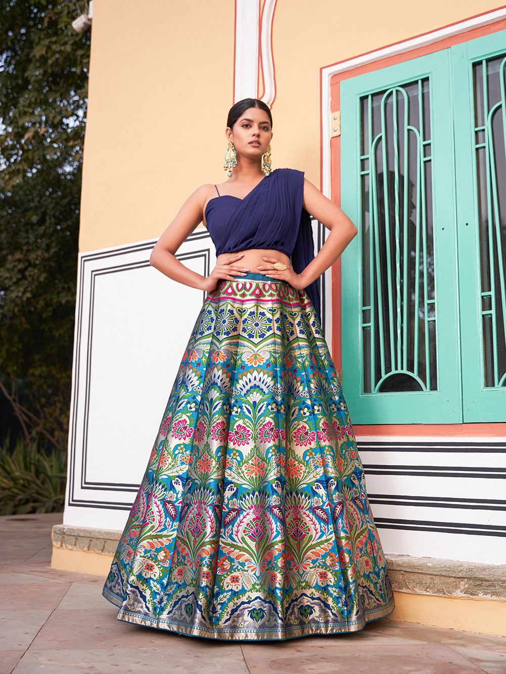 Woman wearing a multicolor banarasi silk lehenga with traditional floral motifs, styled with a modern navy blue one-shoulder blouse – perfect for weddings, Diwali, Rakhi, and festive events in UAE by Meraki Vastram.