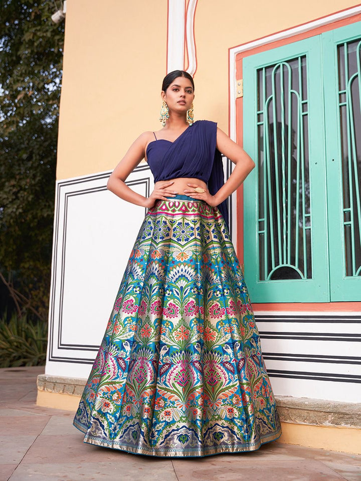 Woman wearing a multicolor banarasi silk lehenga with traditional floral motifs, styled with a modern navy blue one-shoulder blouse – perfect for weddings, Diwali, Rakhi, and festive events in UAE by Meraki Vastram.