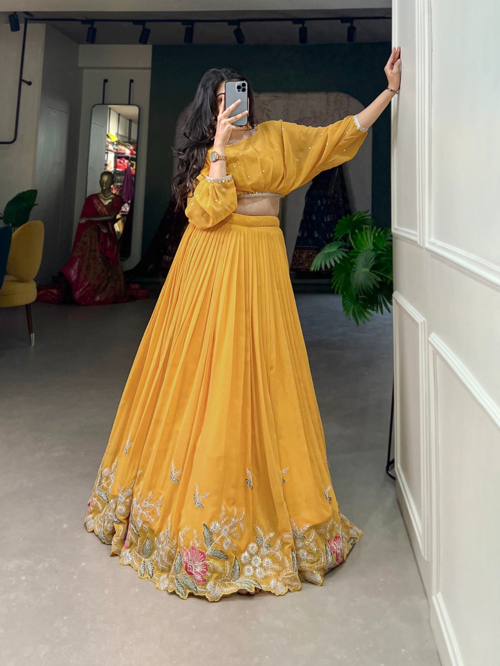 Woman wearing a yellow pleated lehenga with floral embroidery and balloon sleeve blouse taking a mirror selfie- perfect for Haldi, Teej, Diwali, Ganpati and festive celebrations – Meraki Vastram ethnic wear in UAE.